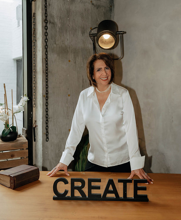 Irene Papadoulis of The Workplace Community Podcast in a white shirt standing behind a wooden table with the word 'CREATE' on it, in an indoor setting.