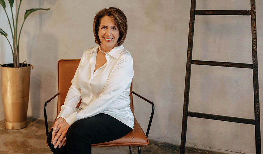 Irene Papadoulis of The Workplace Community Podcast sitting on a chair in a room with a ladder and plant
