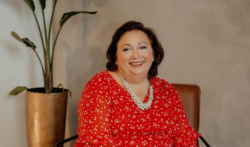 Mary Kelly of The Workplace Community Podcast in a red dress sitting in a chair with a plant in the background