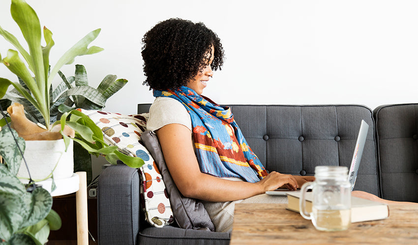 Woman sitting on a couch with a laptop, surrounded by plants in a home setting.