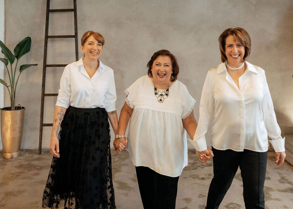 The women of The Workplace Community Podcast holding hands and smiling in a bright, modern room with a textured gray wall, a wooden ladder, and a potted plant. The woman on the left wears a white button-up shirt and a long black lace skirt.